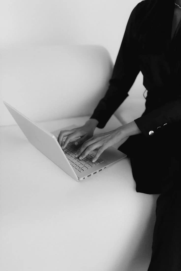 Mãos de uma pessoa vestida de preto digitando em um laptop sobre um sofá branco.